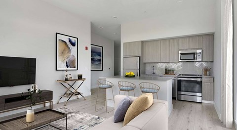 A modern living room with a flat screen TV, a coffee table, and a kitchen area in the background.