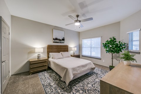 Austin, TX, Apartments Near Texas Department of Public Safety - Canyon Ridge - Bedroom With Nightstands, Lamps, Wall Art, Ceiling Fan, Area Rug, Dresser, Potted Plant, and Large Windows.