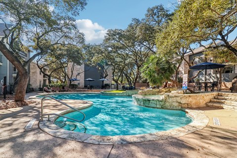 San Antonio, TX, Apartments Near Bowlero Thousand Oaks - Rock Canyon Apartments - Lagoon-Style Pool With Stone Edging, Lounge Chairs, Umbrella Seating, and Mature Trees.