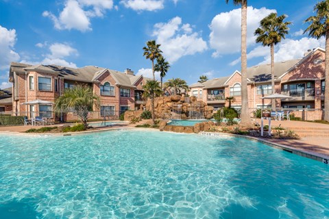 West Houston, TX, Apartments Near Shell Houston - San Montego - Resort-Style Pool With Rock Waterfall, Palm Trees, Lounge Seating, And Brick Apartment Buildings Under Blue Sky.