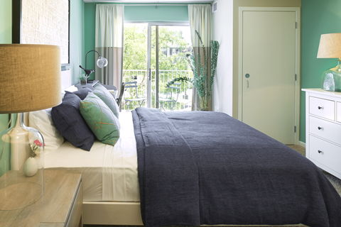 Woodbury, MN, Apartments Near Sky Zone Trampoline Park - The Barrington - Bedroom With Green Accent Walls, A Dark Bedspread, White Dresser, Table Lamps, And Sliding Glass Door to Balcony.