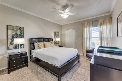 Dog-Friendly Apartments in Austin, TX - The Cliffs at Barton Creek - Bedroom With Dark Wood Bed, Carpet Flooring, Ceiling Fan, Nightstands, Lamps, Dresser, Wall Art, and Large Window.