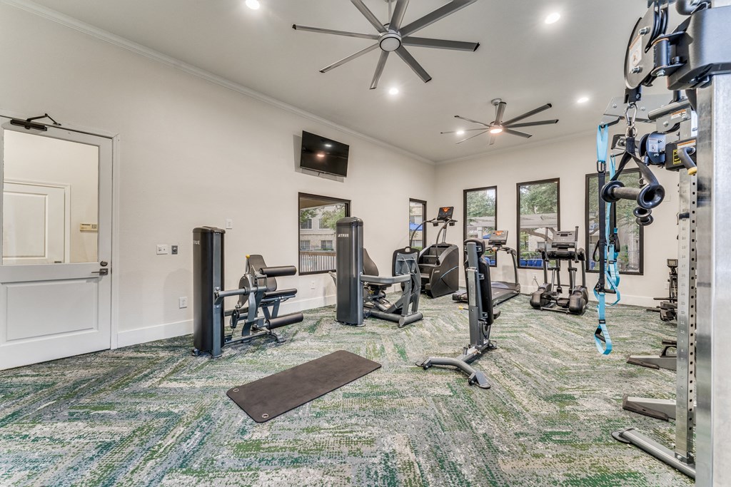 Pet Friendly Apartments in Wells Branch Austin TX - Parkside Crossing - Gym with Carpetted Floor, Cardio Machines, Weightlifting Equipment, Large Mirror, Ceiling Fan, Windows, and TV Screen