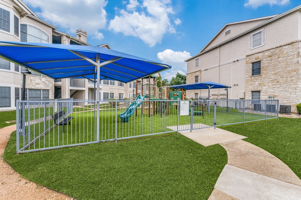 Austin, TX, Apartments - Parkside Crossing - Gated Playground with Manicured Grass, Jungle Gym, and Shaded Bench Seating.
