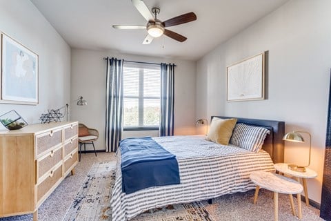 Allen, TX, Apartments With In-Unit Washer and Dryer - Tacara Steiner Ranch - Bedroom With Large Window, Ceiling Fan, Wood Dresser, Nesting Tables, Lamps, and Armchair.