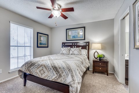 Dog-Friendly Apartments in Austin, TX - The Flats on San Felipe - Bedroom With A Wooden Bed Frame, Patterned Bedding, Carpeted Flooring, and Window Blinds.