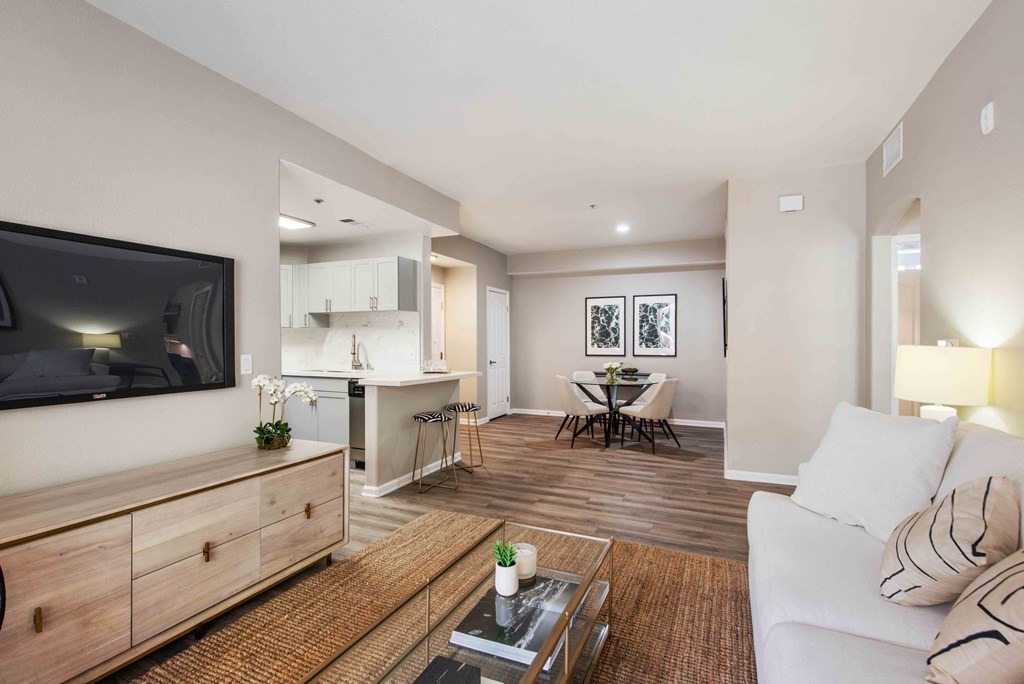 Dog-Friendly Apartments in Camarillo, CA - Allure - Interior Living Room with Wood Style Flooring, an Area Rug, a Wall Mounted TV, a White Couch, a Connected Kitchen Area with a Dishwasher and White Cabinets, and a Dining Area with a Round Glass Table and White Chairs.
