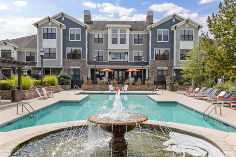 Dog-Friendly Apartments in Charlotte, NC - The Apartments at Blakeney - Swimming Pool with a Water Fountain, Sun Loungers, Lounge Chairs, Small Tables, Umbrellas, and Lush Landscaping.