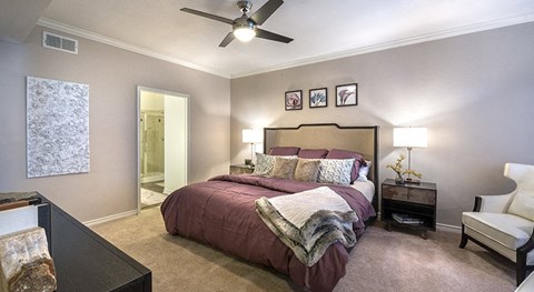 Apartments in McKinney, TX, With In-Unit Washer and Dryer - The Reserve at Stonebridge Ranch - Bedroom With Plush Carpet, Ceiling Fan, Nightstands, Lamps, Accent Chair, and Ensuite Bathroom Entry.