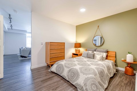Studio Apartments In Green Lake, Seattle, WA - Circa Green Lake - Spacious Bedroom With Hardwood Floor, Bed, Round Mirror, Nightstand with Lamp, Round Side Table, Dresser, and View of Living Room.