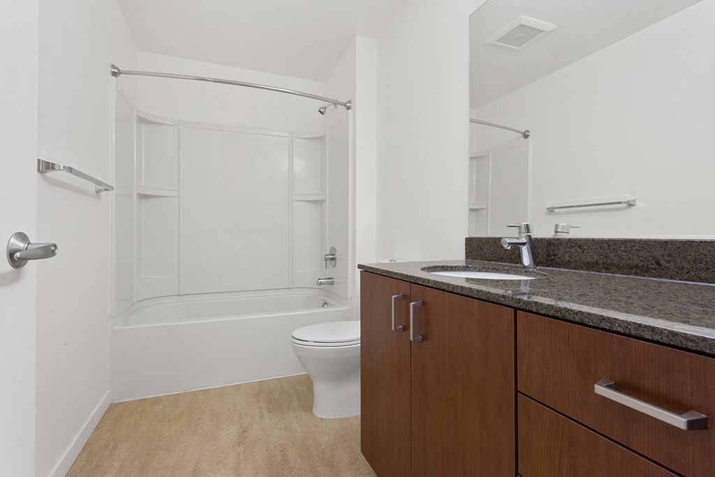 Apartments Nob Hill, Portland, OR - The Cordelia - Bathroom with Granite Countertops, Wood-Style Vinyl Flooring, Brown Cabinets, Large Mirror, and Shower and Bathtub Combo.