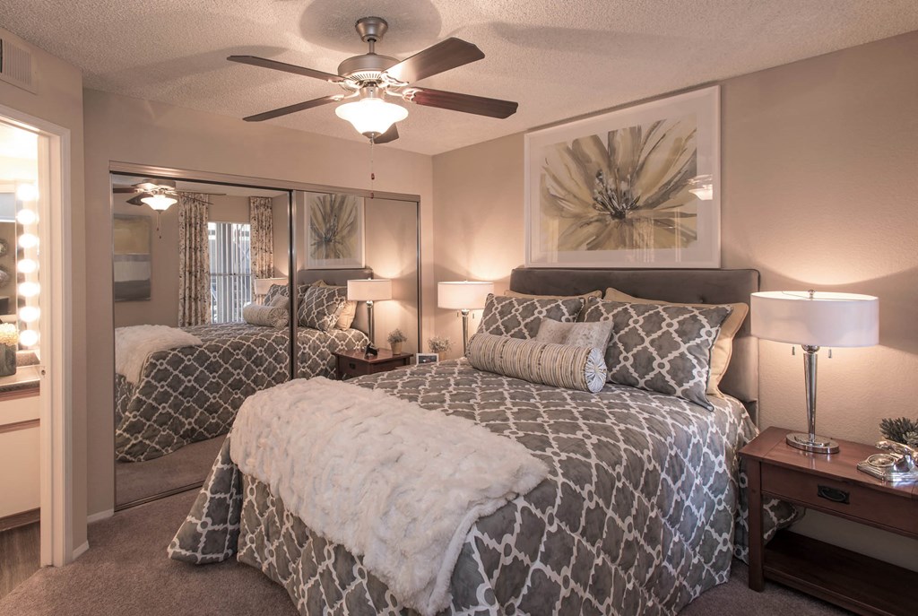One and Two-Bedroom Apartments in Ahwatukee, AZ - Pacific Bay Club - Bedroom with Mirrored Closet Doors, Carpet Floors, Ceiling Fan, Cozy Bed, Stylish Decor, and Door to Bathroom with Wood-Style Plank