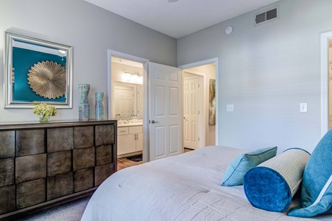 Woodstock, GA, Apartments Near Cherokee County Government - Lea Woodstock - Bedroom With A White Bed, Blue Accent Pillows, Dark Wood Dresser, Wall Art, And Attached Bathroom.