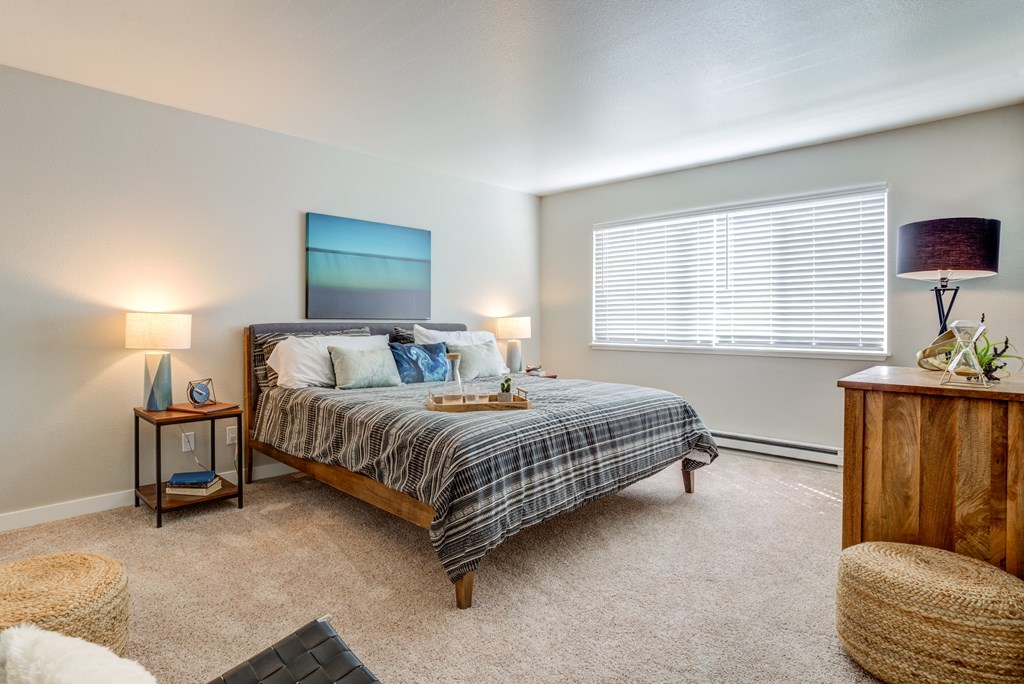 One-Bedroom Apartments in Mountain Park, OR - MiLO at Mountain Park - Bedroom with Carpeting, Large Window, Wooden Dresser, and 2 Nighstands with Lamps