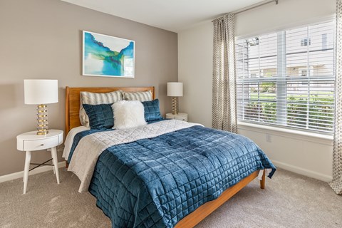One-Bedroom Apartments in Marlborough, MA - Orchards Apartments - Bedroom With A Wooden Bed, Blue Quilted Bedding, White Nightstands, Table Lamps, and Patterned Curtain.