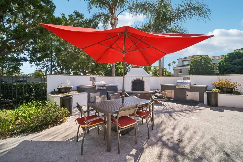 Camarillo, CA Pet-Friendly Apartments - Allure - Outdoor BBQ Picnic Area with Two Grilling Stations, a Fireplace, a Table with a Large Red Umbrella and Surrounding Chairs.