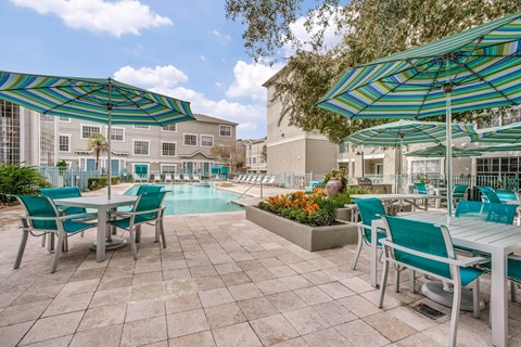 2-Bedroom Luxury Apartments in Montrose, Houston, TX - The Maroneal - Pool Surrounded by the Apartment View, Featuring a Lounge Area, Patio with Stone Pavers, Tables and Mesh Chairs, and Umbrellas.