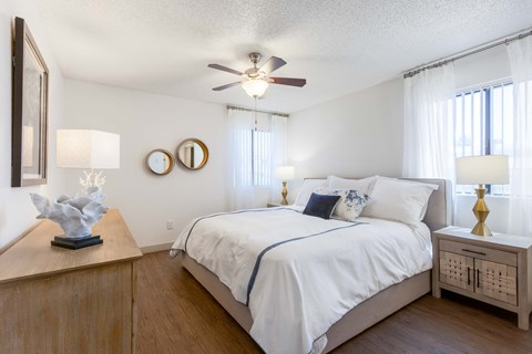 Scottsdale, AZ Apartments for Rent - The Sycamore at Scottsdale - Bedroom with Hardwood-Style Flooring, Bed with White Bedding and a Cushion Bedframe, Nightstands with Lamps, Large Wooden Dresser, Ceiling Fan with a Light, and Two Windows with White Blinds and Curtains.