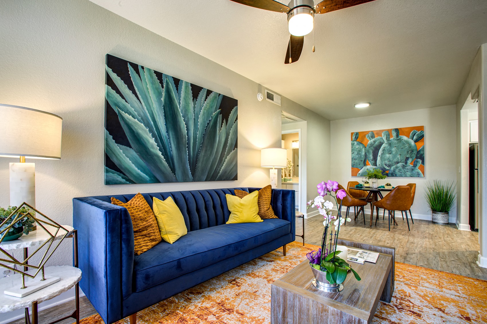 South Phoenix Apartments for Rent - Village at Lakewood - Living Room with Wood-Style Flooring, Orange Area Rug, Blue Sofa, Wooden Coffee Table, Ceiling Fan, and Wall Art