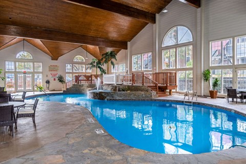 Omaha, NE, Apartments With Attached Garage - Apple Creek Apartments - Indoor Pool With Curved Swim Area, Stone Fountain, Wood Beam Ceiling, Large Windows, and Poolside Seating.
