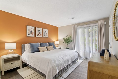 Two-Bedroom Apartments in Marietta, GA - Wood Pointe Apartment Homes - Bedroom With Wood-Style Plank Flooring, Orange Accent Wall, White Bedding, Nightstands, Sliding Glass Door, And Wood Dresser.