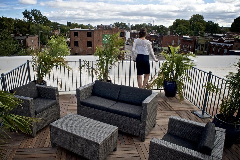 A woman in a white blouse and black skirt stands on a balcony with outdoor furniture.