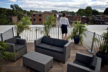 A woman in a white blouse and black skirt stands on a balcony with outdoor furniture.