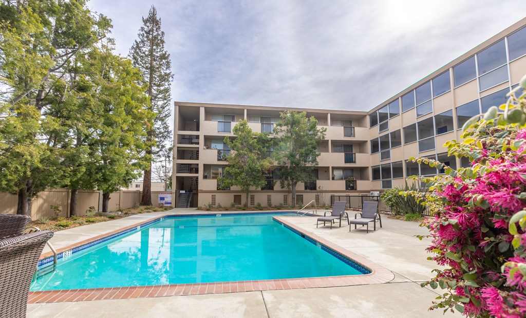 A swimming pool is surrounded by trees and chairs in front of a building.
