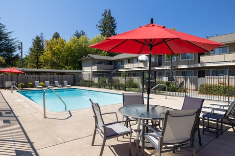 A red umbrella shades a table with chairs around a pool.