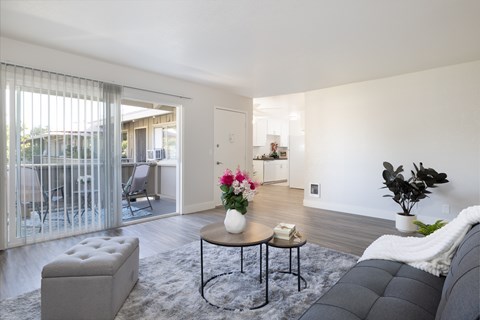 A modern living room with a grey sofa, a round table with a vase of flowers, and a large window with blinds.