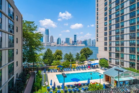 A swimming pool with blue umbrellas and chairs is surrounded by buildings.