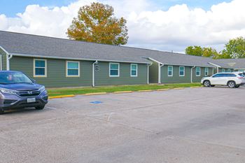 A parking lot with a blue car and a white car parked in front of a building with green siding.