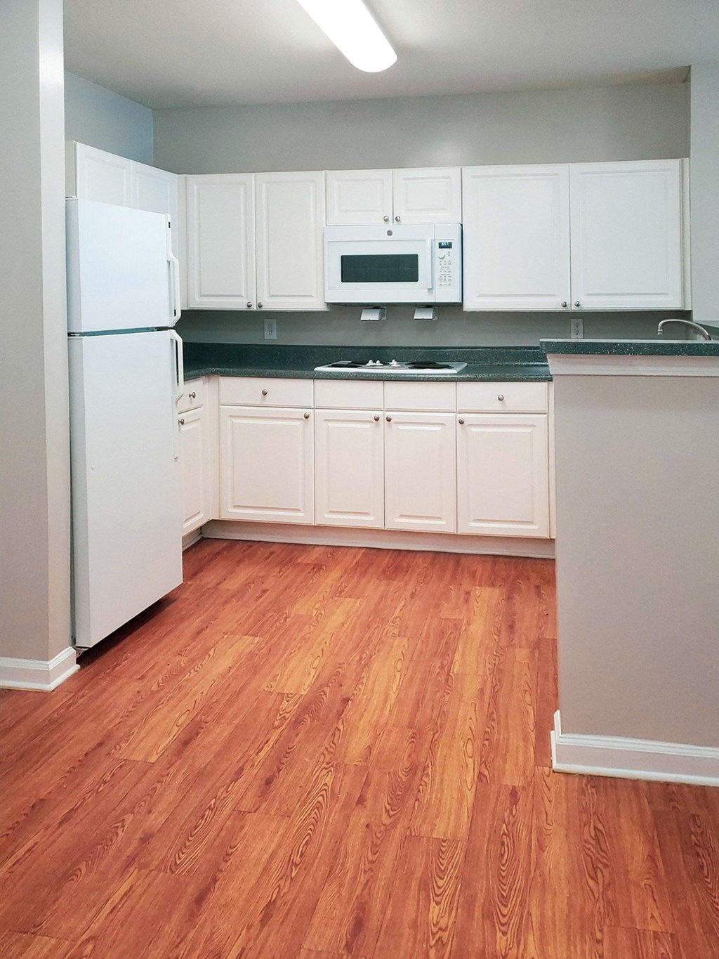 A kitchen with white cabinets, dark countertops, and wood flooring. It includes a white refrigerator, microwave, and an electric stovetop. A raised breakfast bar partially separates the space
