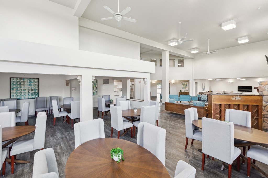 Spacious community dining and lounge area featuring round wooden tables with high-back upholstered chairs, a high ceiling with ceiling fans, and modern decor. The open-concept space includes a cozy seating area, a fireplace, and a kitchen in the background, creating a welcoming atmosphere