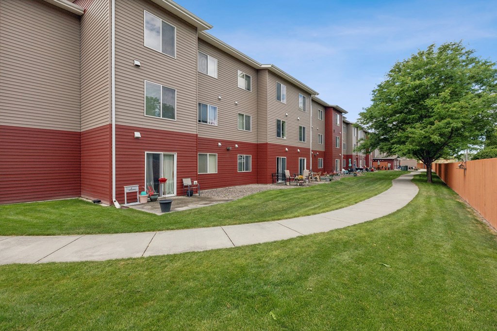 Three-story apartment building with beige and red siding, featuring large windows and patio doors on the ground level. A curved walking path winds through the well-manicured lawn, lined with mature trees and a wooden privacy fence