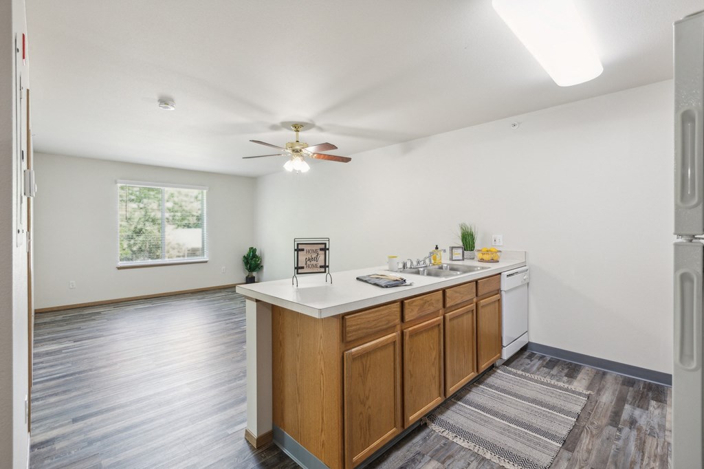 Open-concept apartment kitchen and living area featuring wood cabinetry, a white countertop, and a dishwasher. The space has wood-style flooring, a ceiling fan with a light fixture, and a large window providing natural light