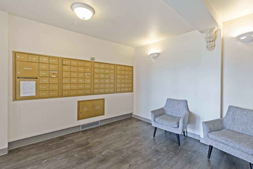 Apartment mailroom featuring a wall of gold-colored mailboxes, two gray upholstered chairs, and warm lighting. The space has wood-style flooring and a neutral color palette, creating a clean and inviting environment