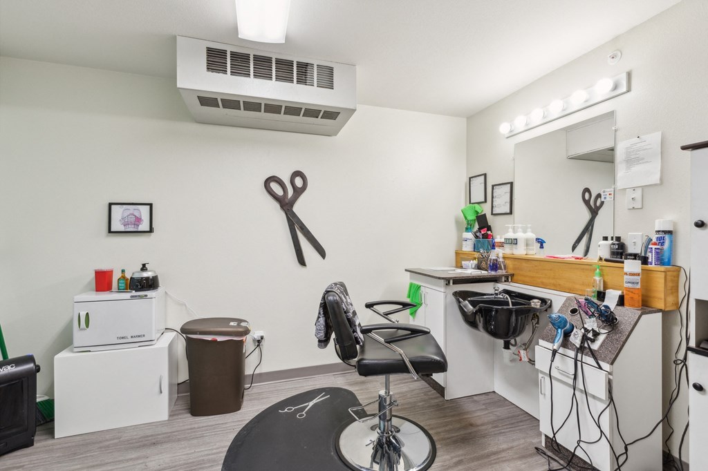 Community hair salon featuring a black styling chair, a wash station with a sink, and a well-lit vanity mirror. The space includes salon tools, hair care products, and decorative elements such as wall-mounted scissors, creating a functional and inviting setting