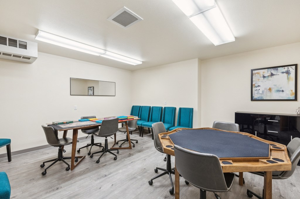 Community game and activity room featuring a poker table with cup holders, a long table with board games, and teal upholstered chairs along the wall. The space has wood-style flooring, bright overhead lighting, and modern decor for a welcoming atmosphere