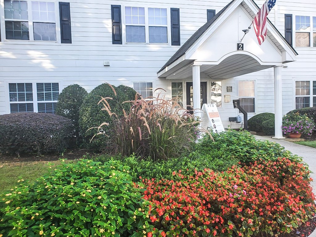 The entrance of a white residential building with black shutters and a gabled awning displaying an American flag. The surrounding landscaping includes neatly trimmed bushes, ornamental grasses, and colorful flowers. A walkway leads to the entrance, where a sign and mailbox are visible