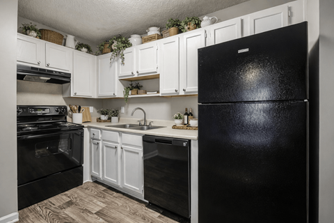 A black fridge stands in a kitchen with white cabinets and a wooden floor.