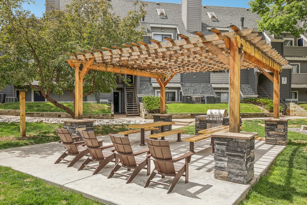 A wooden pergola with chairs and a table is in the foreground of a residential area.