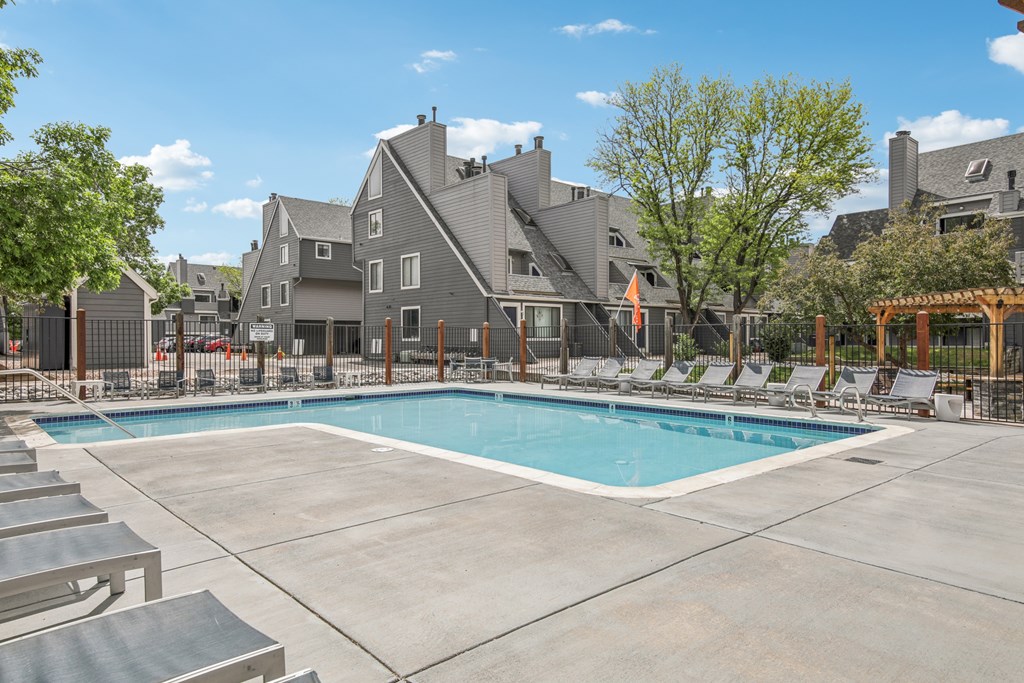 A large swimming pool surrounded by lounge chairs and a fence with a building in the background.