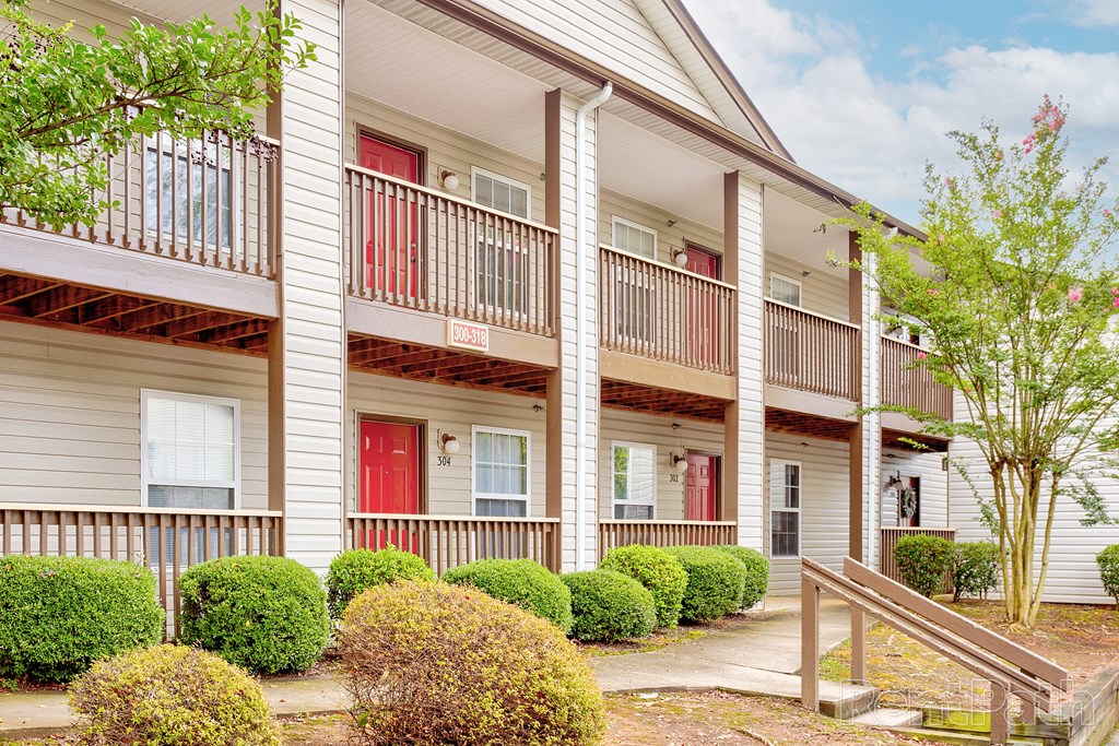 Exterior view of a two-story apartment building featuring beige siding, brown wooden railings, and red front doors. The units have private balconies on the upper level and small porches on the lower level. Lush green shrubs and a well-maintained walkway add to the inviting atmosphere, with a clear blue sky and light clouds in the background
