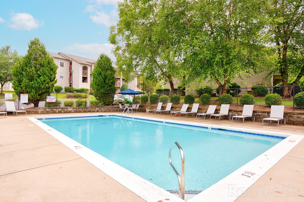 Outdoor swimming pool in a residential community, surrounded by lounge chairs and lush greenery. The pool area is enclosed by a black metal fence with trimmed bushes, and multi-story apartment buildings with balconies are visible in the background. The water is clear and inviting under a bright sky with scattered clouds