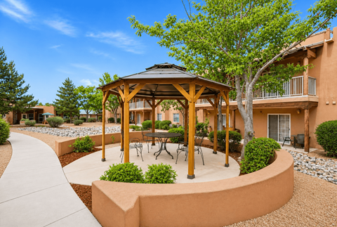 A gazebo with a table and chairs is surrounded by bushes and trees.