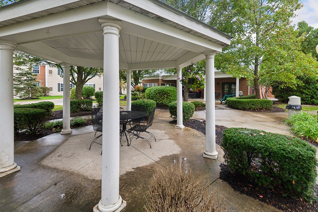 A white gazebo with a table and chairs is surrounded by columns and bushes.