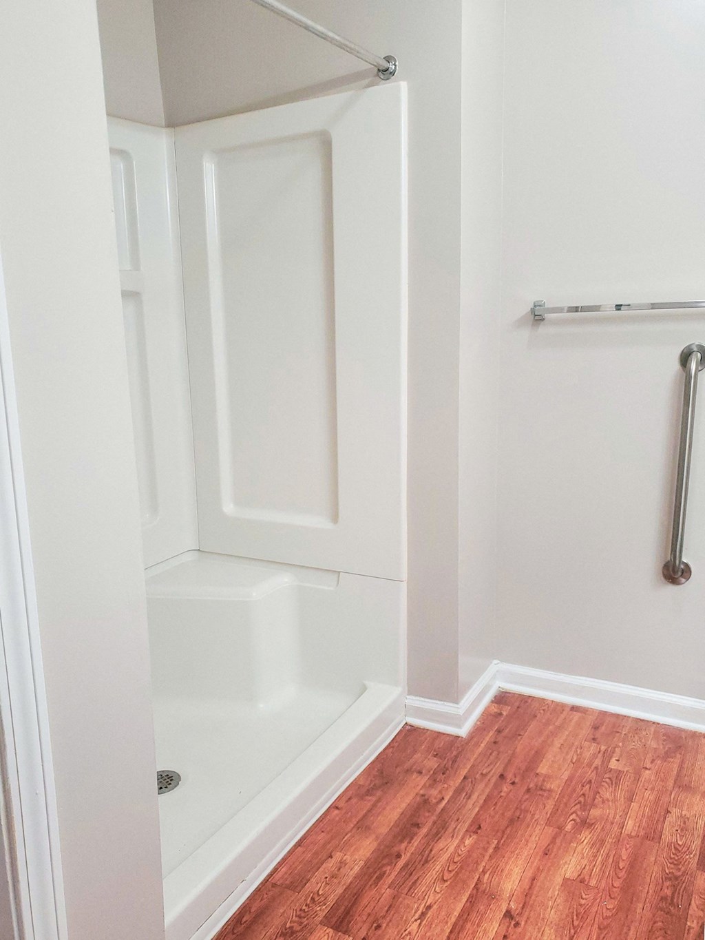 A bathroom with a white walk-in shower that has built-in seating and a shower rod. The walls are light-colored, and the floor has wood-style flooring. A towel rack and a grab bar are mounted on the wall