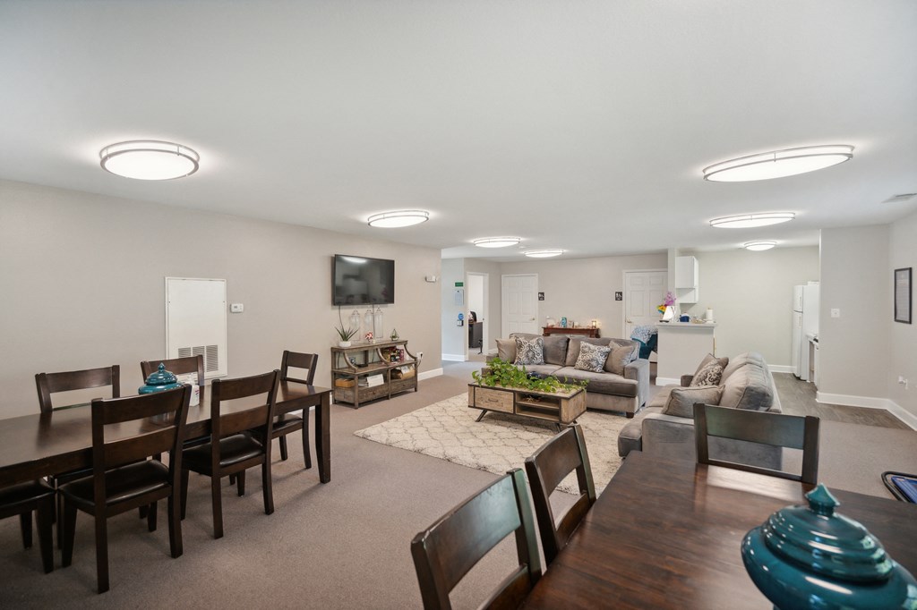 Large, inviting community space featuring dining tables with wooden chairs, comfortable sofas arranged around a coffee table, a wall-mounted television, and modern overhead lighting.