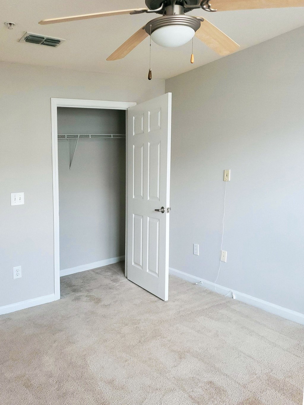 An empty bedroom with beige carpeting, light gray walls, and a ceiling fan with a light. An open door reveals a small closet with a wire shelf
